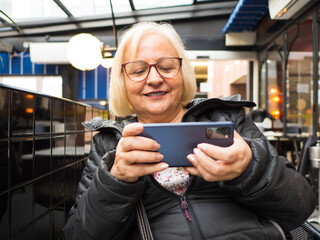blonde gamer grandmother with glasses playing on the mobile on a restaurant terrace