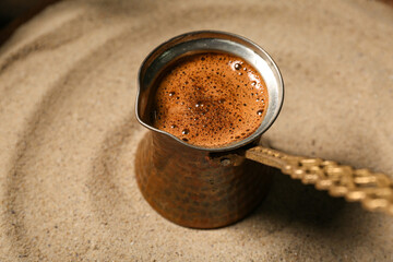 Cezve of hot coffee on sand background
