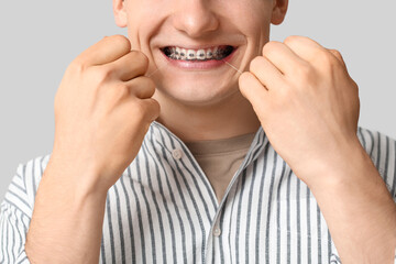 Young man with dental braces flossing teeth on light background, closeup