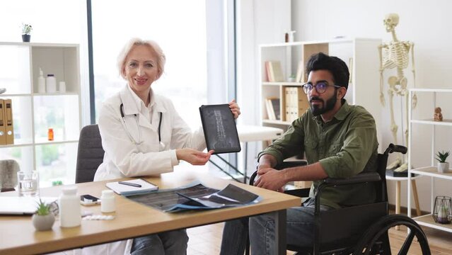 Portrait of senior female doctor and bearded male patient with disability posing at office desk in clinic. Health professional giving recommendation about treatment due to CT scan images.