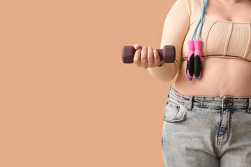 Overweight woman with dumbbells and jumping rope on beige background. Weight loss concept