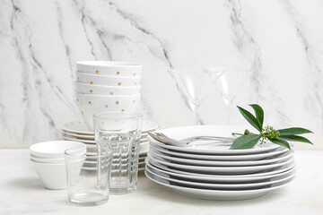 Composition with clean dishes, cutlery, glasses and leaves on white table near marble wall