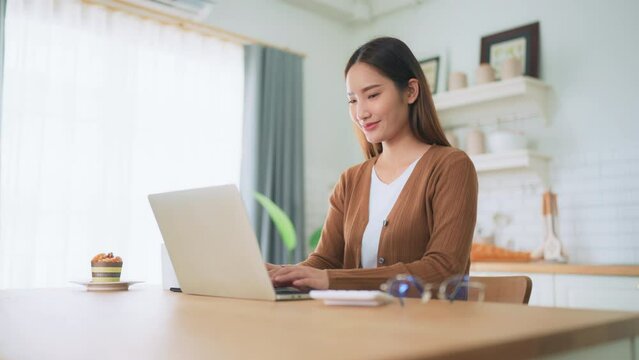 Beautiful young Asian woman working with laptop computer in home kitchen. Work at home