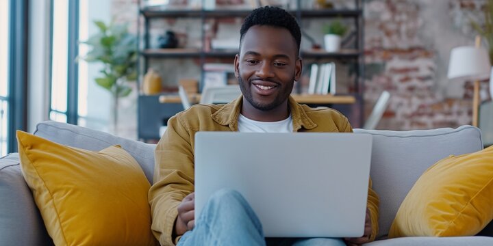 A handsome black African American man is working on a laptop computer while sitting on a sofa in a cozy living room. Freelancer working from home. Browsing Internet, Using Social Networks,
