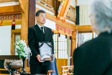 お葬式で遺影を持って挨拶する喪主の高齢者男性（喪主の挨拶）
