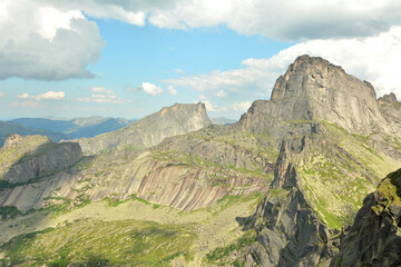 High and narrow ridges of jagged mountain peaks, partly covered with grass under a cloudy summer sky.