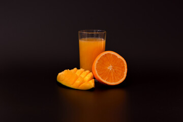A glass glass of a mixture of tropical fruit juice on a black background, next to pieces of mango and ripe orange.