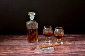 A crystal decanter and two glasses of brandy on a dark wooden table, next to a glass ashtray with a Cuban cigar.