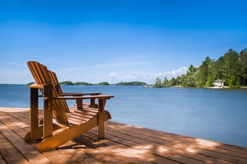 Two Adirondack chairs grace a wooden dock on a sunny summer morning in Muskoka, Ontario, Canada,...