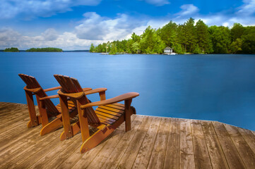 Two Adirondack chairs rest peacefully on a wooden dock, gazing out over the serene, blue lake. A...