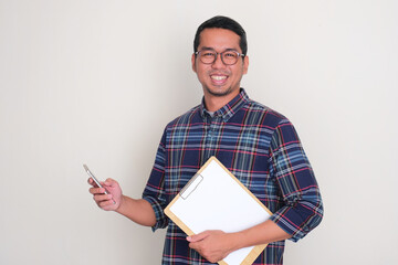 A man smiling at the camera while holding mobile phone and paper on clipboard
