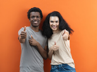 Happy Couple Giving Thumbs Up Sign, Young Adults Approval Gesture, Positive Feedback, Satisfaction Concept, Interracial Relationship, Joyful Expression, Casual Attire, Orange Background