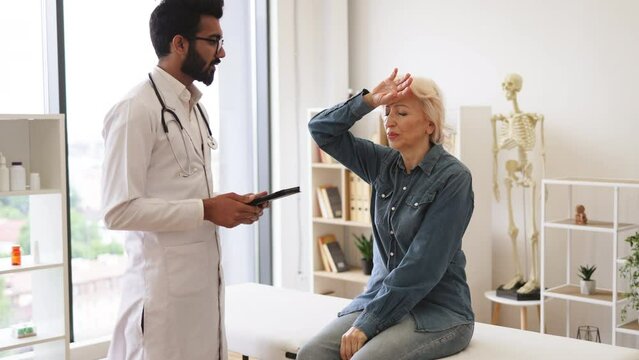 Old woman describing symptoms of illness to young male doctor at moderm medical office. Adult bearded physician giving recommendations and using tablet for writing electronic prescription.
