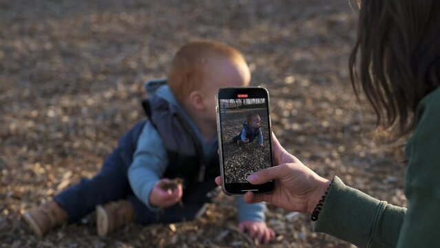 in the playground, one year old red hair  boy sits on the ground and plays with the ground, explores nature, the mother takes photos and videos on the phone for childhood memories