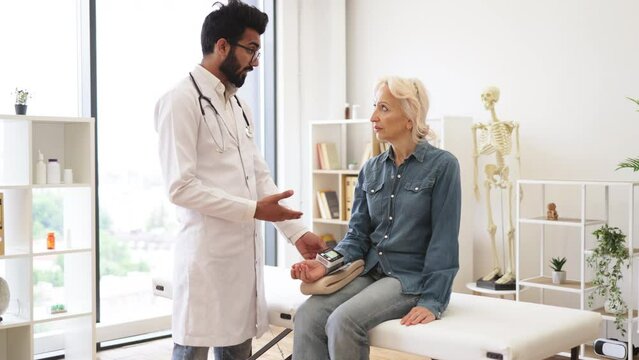 Bearded young male cardiologist telling elderly female patient about all possible reasons of hypertension. Adult woman listening explanations about necessity of leading healthy lifestyle.