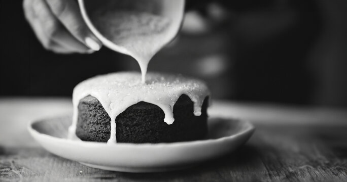 A baker delicately pours a thick, sweet frosting onto a small layer cake in shallow depth of field.
