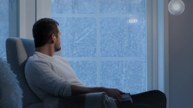 Pensive man sitting in armchair near window at evening looking at snowfall. Static