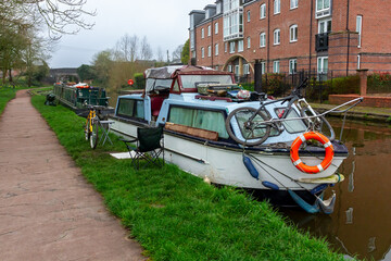 A canal house boat is moored by the towpath. There are bicycles parked outside. One is on the grass...