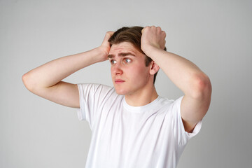 Young Man In White T-Shirt Looking Worried With Hands On Head Against Plain Background