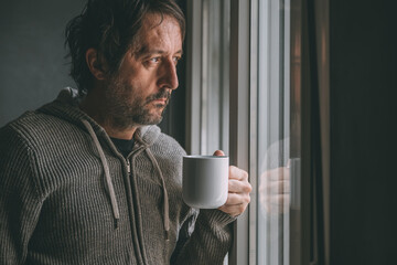 Pensive adult male drinking coffee in the morning by the apartment window while looking out and thinking. Caffeine dose for starting of the day.