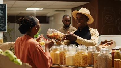 Farmer with apron recommending new homemade sauce for woman, showing organic pasta types and other pantry supplies. African american vendor presenting products to regular customer. Camera 1.
