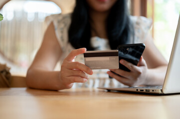 An Asian woman holding a credit card and a smartphone while sitting at a table in a cafe.