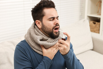 Young man with scarf using throat spray indoors - obrazy, fototapety, plakaty