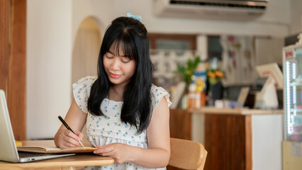 A beautiful Asian woman focusing on writing something in a notebook while sitting in a coffee shop.