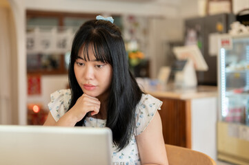 A serious, thoughtful young Asian woman is focusing on her work, working remotely at a coffee shop.