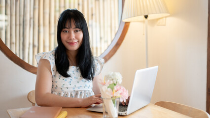 A cute Asian woman sits at a table in a coffee shop with her laptop computer, smiling at the camera.