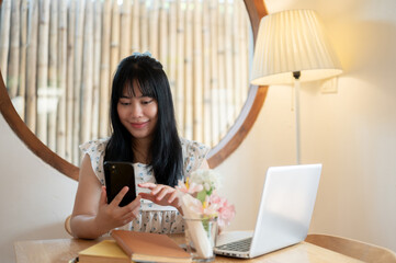 A happy Asian woman is reading messages, using her smartphone while sitting in a coffee shop.