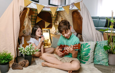 Happy children playing a ukulele and singing on handmade shelter tent in living room. Boy and girl having fun in a cozy diy teepee in house. Staycation and vacation camping at home concept.