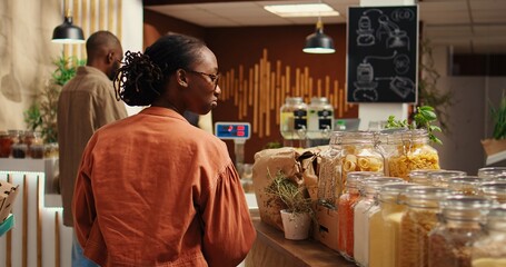 African american client examines bio homemade bulk products, looking to buy natural ethically sourced food alternatives for healthy nutrition. Woman shopping for goods at zero waste store. Camera 2.
