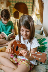 Portrait of smiling little girl playing ukulele while boy reading book on handmade teepee. Children having fun in cozy diy shelter tent in their house. Vacation camping at home or staycation concept.