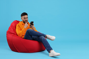 Handsome young man using smartphone on bean bag chair against light blue background. Space for text