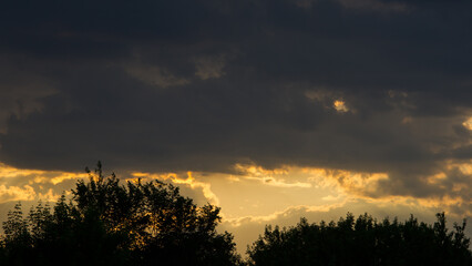 Dark clouds cover the horizon at sunset and dark silhouettes of trees.