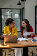 Fototapeta premium A businesswoman in a yellow blazer engaging in a productive discussion with a colleague in a red jacket over work documents in an office.