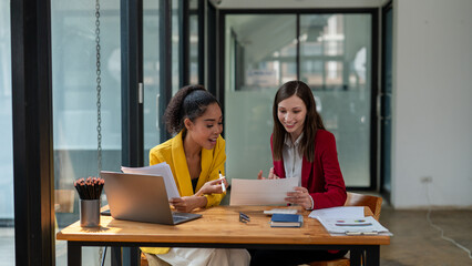 A businesswoman in a yellow blazer engaging in a productive discussion with a colleague in a red jacket over work documents in an office.