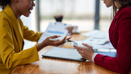 Close-up of a businesswoman’s hands reviewing a detailed bar graph report, with another professional discussing findings in the background.