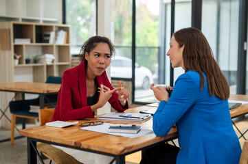 Two professional women in a lively discussion at an office desk, with one in a yellow blazer gesturing and the other showing surprise.