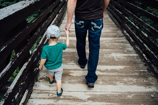 Overhead rear view of dad leading son across bridge in summer.
