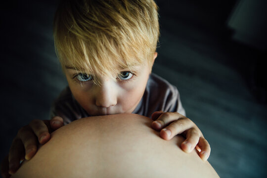 Portrait of small boy kissing moms pregnant baby belly indoors
