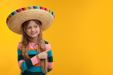 Cute girl in Mexican sombrero hat pointing at something on orange background. Space for text