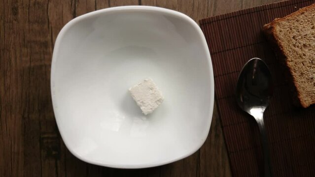 Top view of soft feta cheese cube placed into white bowl with the kitchen knife . 4K