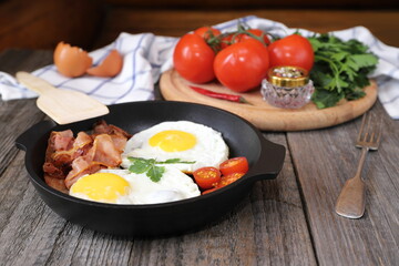 On a wooden table is a pan with fried eggs, tomatoes, bacon and greens.