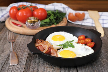 On a wooden table is a pan with fried eggs, tomatoes, bacon and greens.