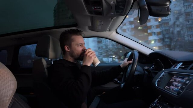 Man enjoying in driving a car on city street in evening, drinking a coffee