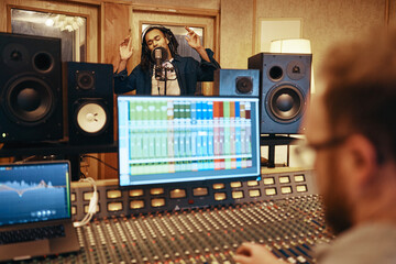 Producer adjusting sound levels during a studio recording session. A black charismatic singer stands and sings with arms raised and eyes closed, into a microphone