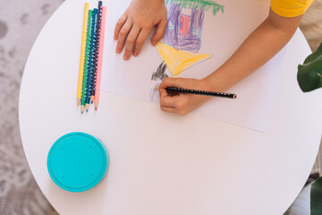 A child draws at the table with a black pencil on a sheet of paper.