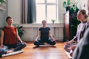 A group of women sit on a cushion on a floor. They sit still with their eyes closed and meditate in a circle during yoga class.